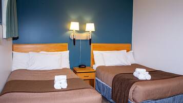 Habitación de hotel con dos camas, cada una con cubrecama marrón y almohadas blancas, un cabecero de madera, una mesita de noche con lámpara y una pared azul.