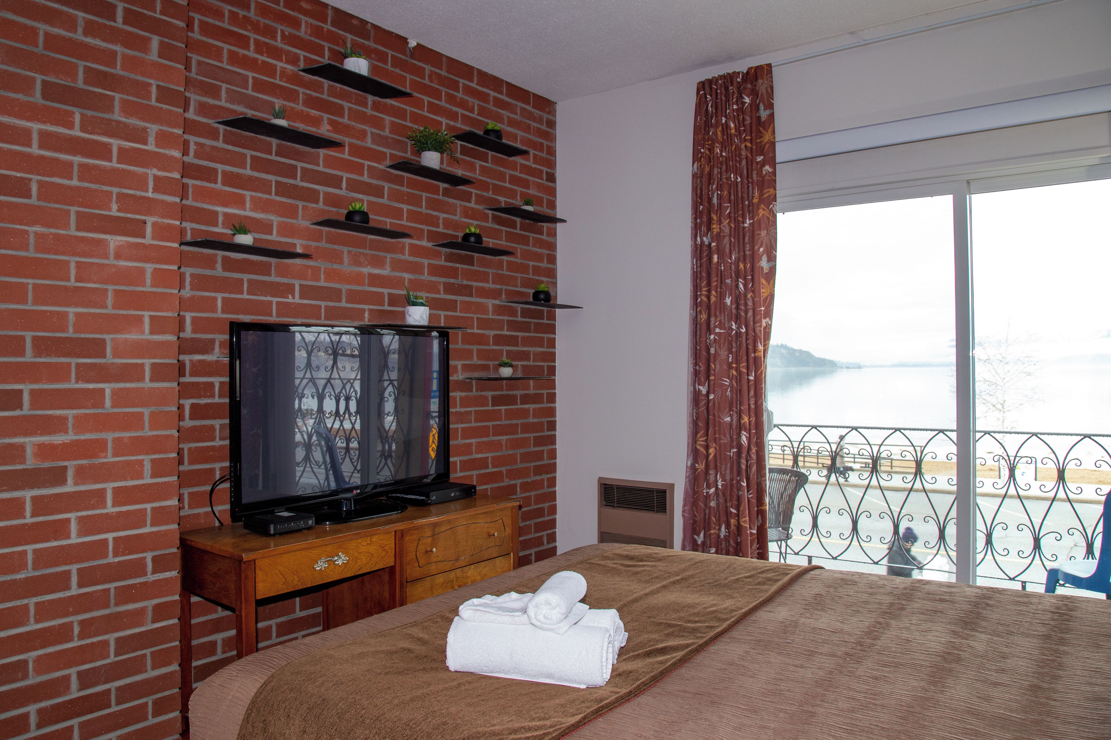 Habitación de hotel con pared de ladrillo, televisor de pantalla plana, cómoda de madera, cama con manta marrón y balcón con vistas.