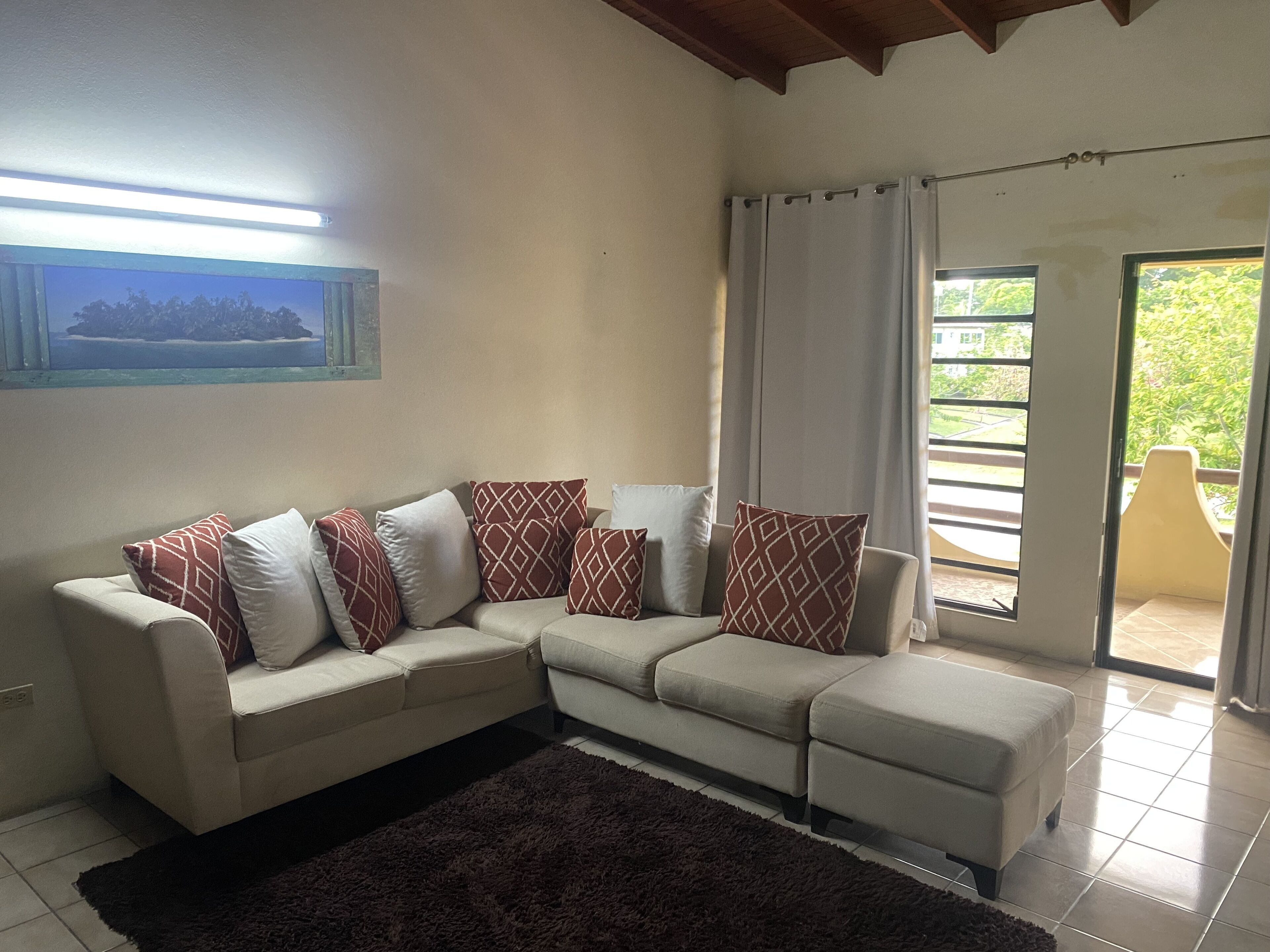 Sala de estar com sofá seccional bege, almofadas decorativas estampadas, tapete escuro e vista para o verde através de uma porta de vidro deslizante.