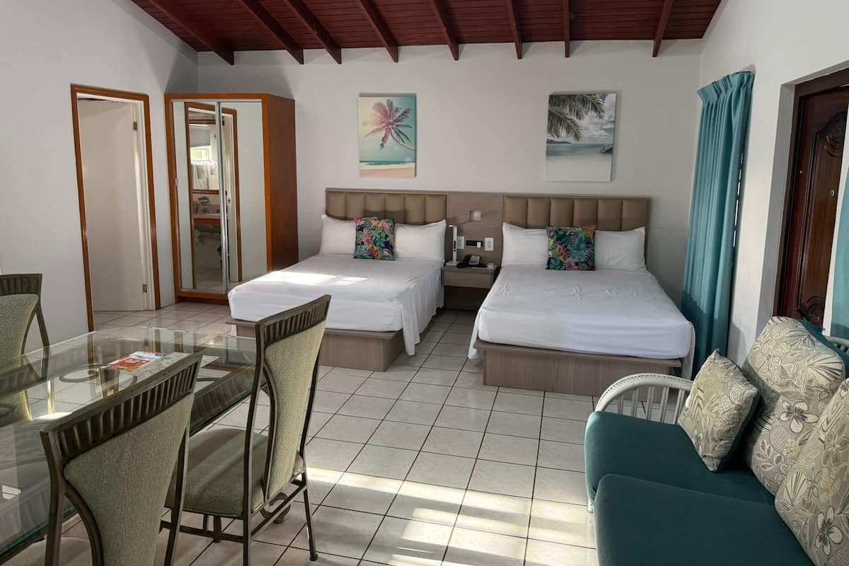 A hotel room with two beds, a glass dining table, chairs, a sofa, and a ceiling with wooden beams.