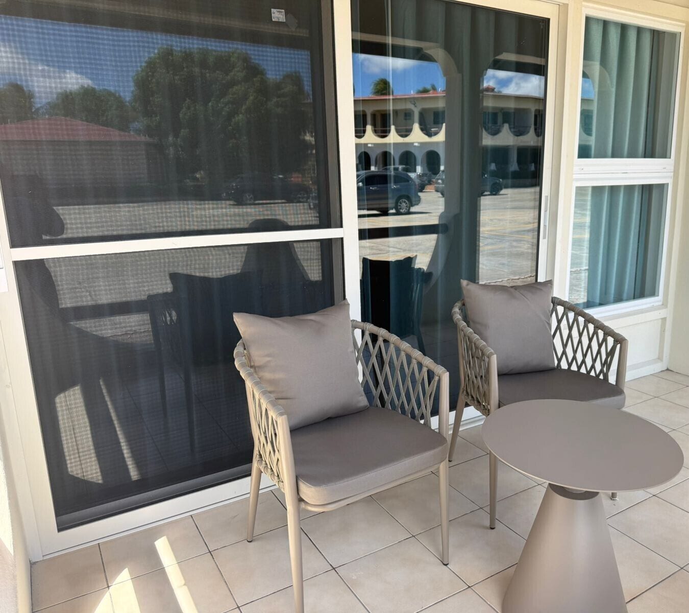 En terrasse med to fletstole og et rundt bord, store vinduer med udsigt til parkerede biler og bygninger, og et flisegulv.