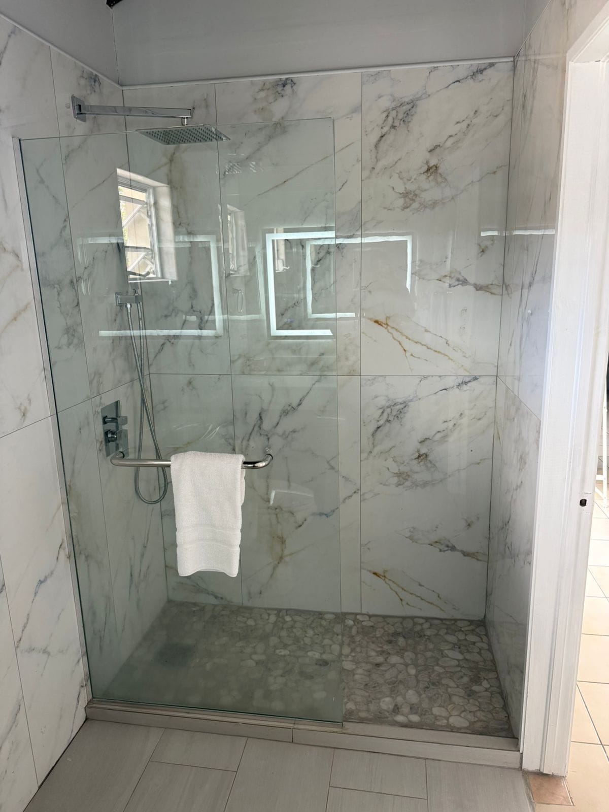 A glass-enclosed shower with marble walls and a white towel hanging on a metal rack.