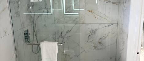 A glass-enclosed shower with marble walls and a white towel hanging on a metal rack.