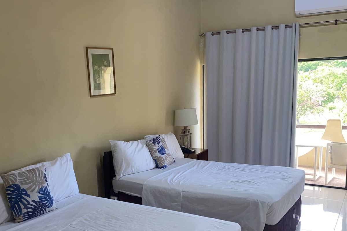 A hotel room with two beds, a ceiling fan, a window with curtains, and a view of greenery outside.