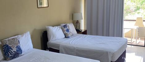 A hotel room with two beds, a ceiling fan, a window with curtains, and a view of greenery outside.