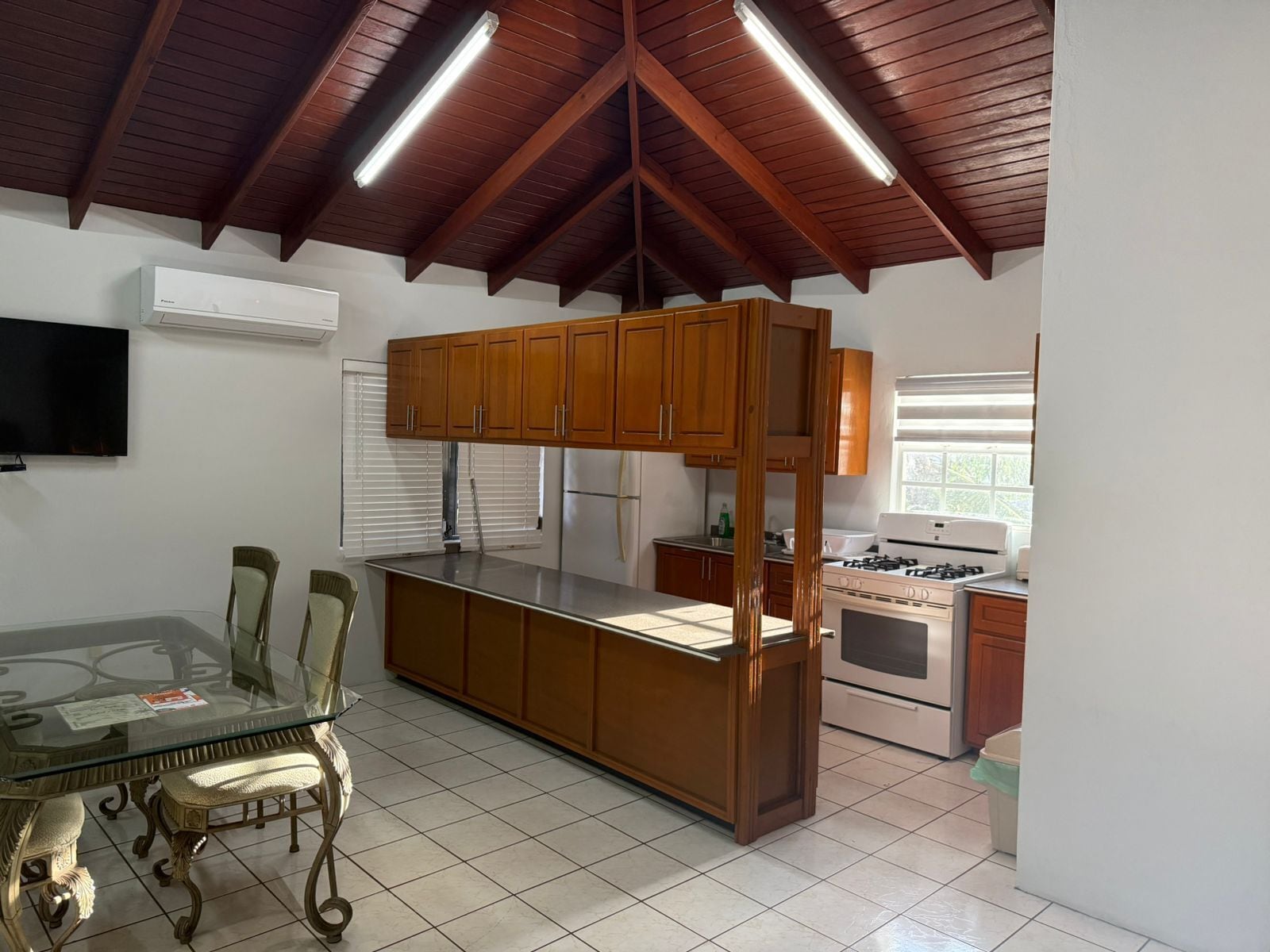 Una cocina con armarios de madera, una mesa de comedor con superficie de vidrio, sillas, estufa y televisor montado en la pared.