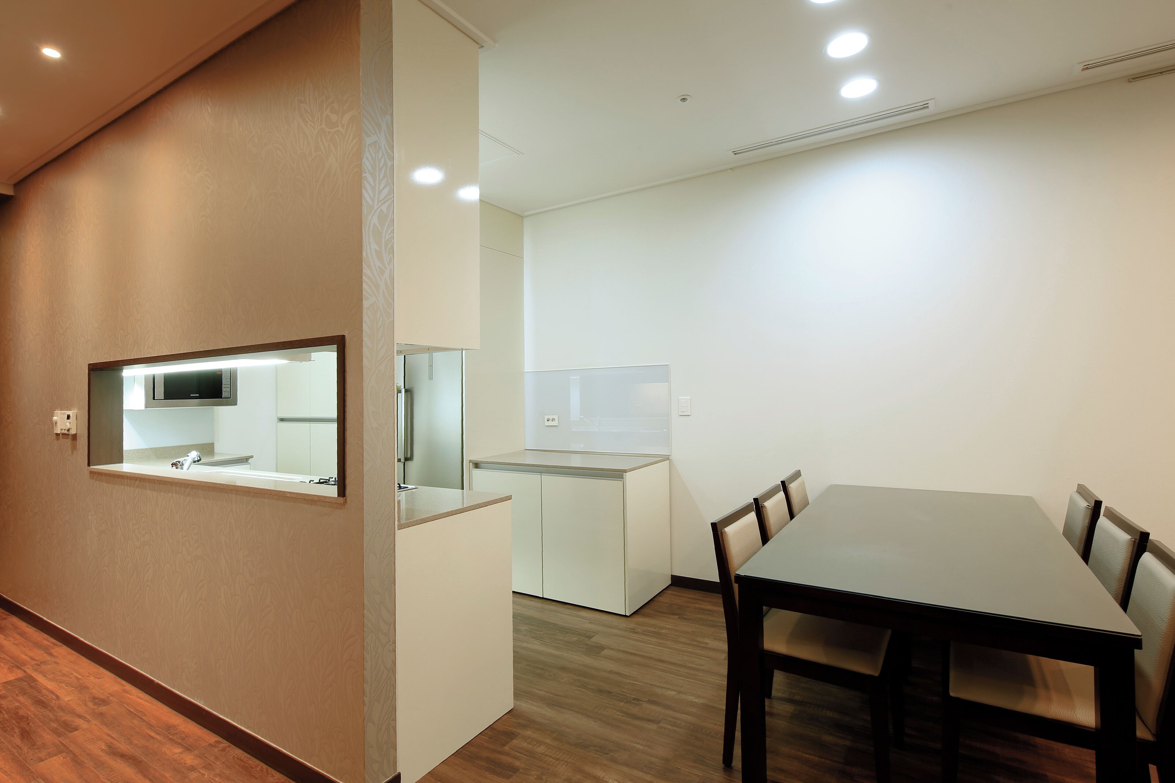 A modern hotel room with a wooden floor, a dining table with chairs, a kitchen area with a microwave, and a large mirror.