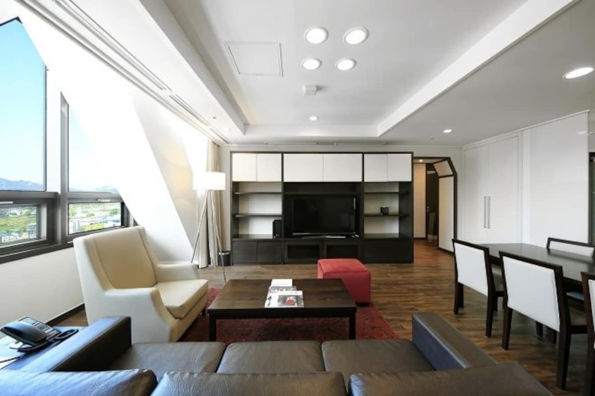 Una sala de estar moderna con un sofá, una mesa de centro y una televisión. Hay un comedor con una mesa y sillas. La habitación tiene un ventanal con vistas al exterior.