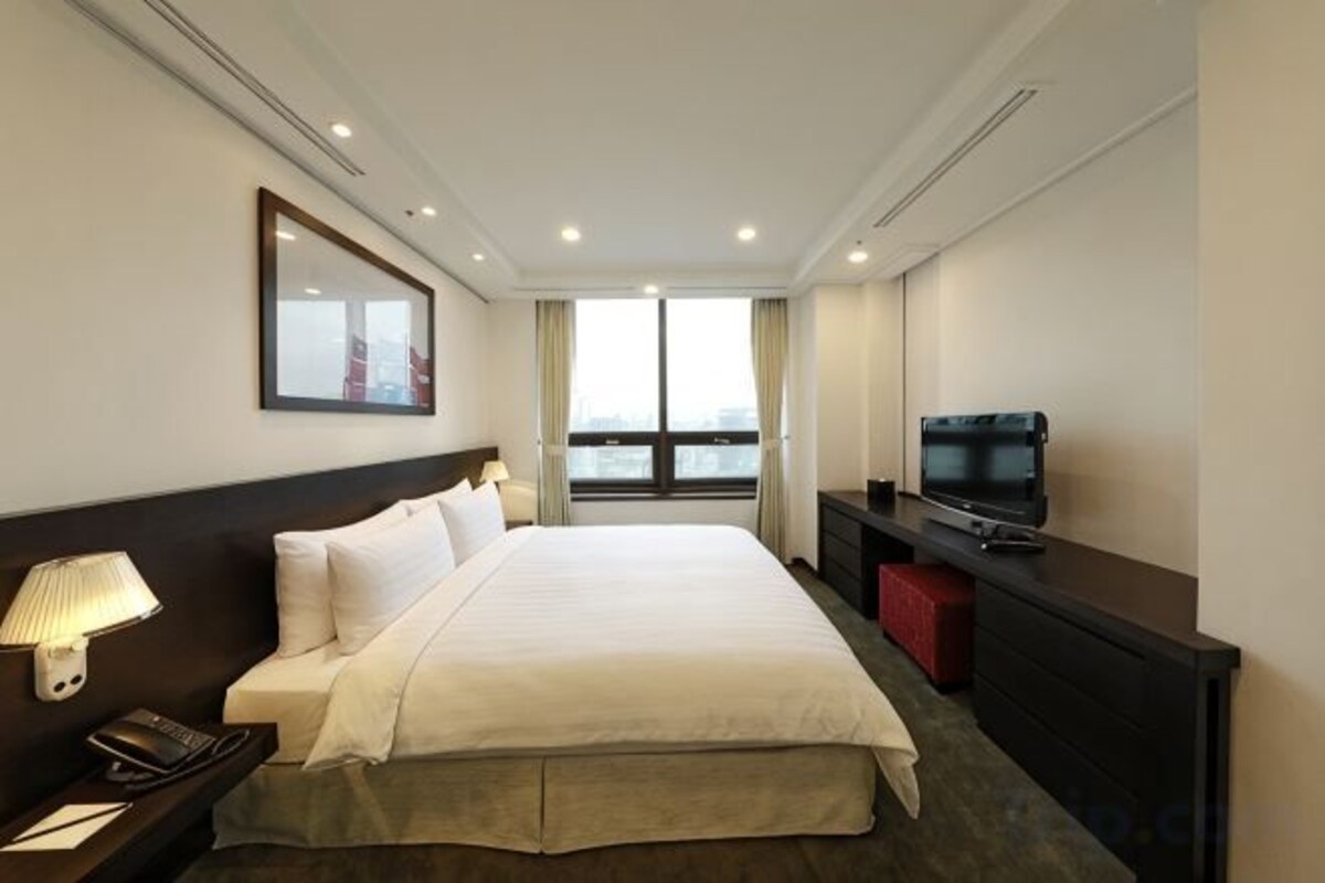 Habitación de hotel con una cama grande, televisión, escritorio con lámpara y vistas al exterior.
