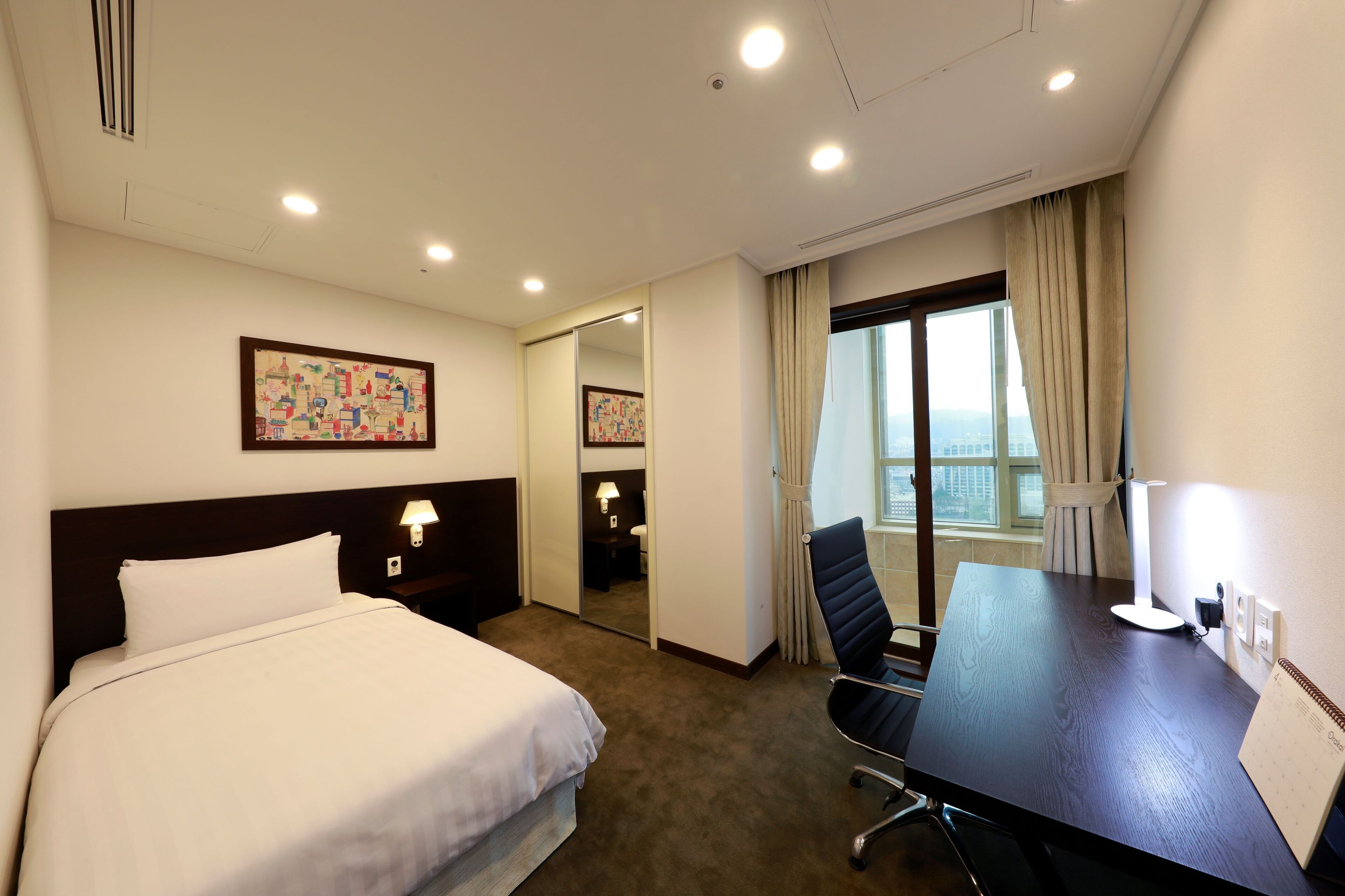 A hotel room with a bed, a desk, a chair, a lamp, a window with curtains, and a painting on the wall.