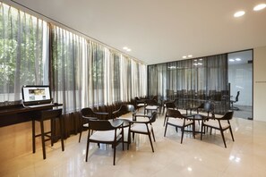 A modern boardroom with a laptop on a desk, large windows with sheer curtains, and a seating area with chairs and tables.