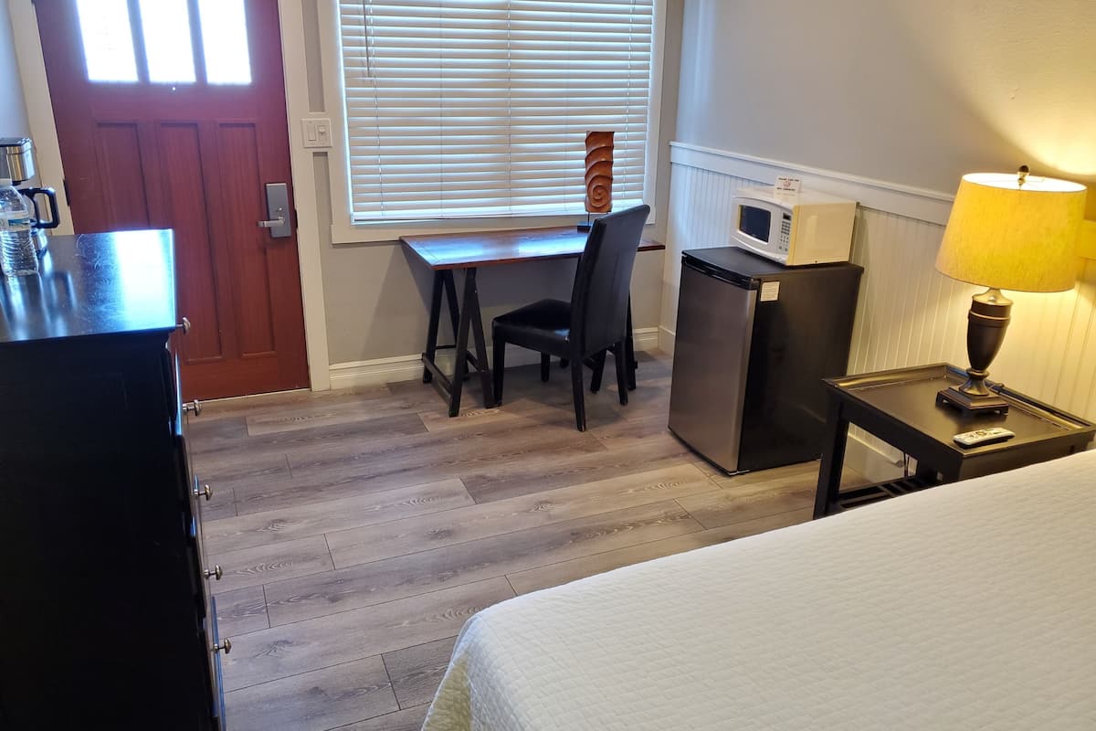 A hotel room with a bed, a desk with a chair, a refrigerator, a microwave, a lamp, and a door with windows.