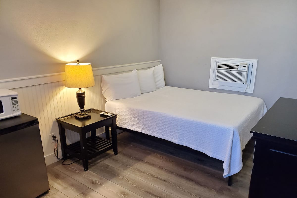 A hotel room with a bed, a bedside table with a lamp, a microwave, a small refrigerator, and an air conditioning unit.