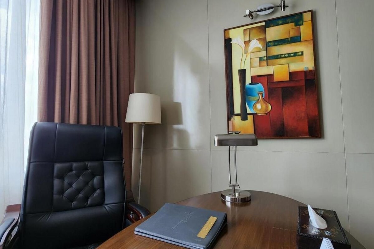 Habitación de hotel con un escritorio de madera, una silla de cuero negro, una lámpara, un cuadro y una ventana con cortinas.