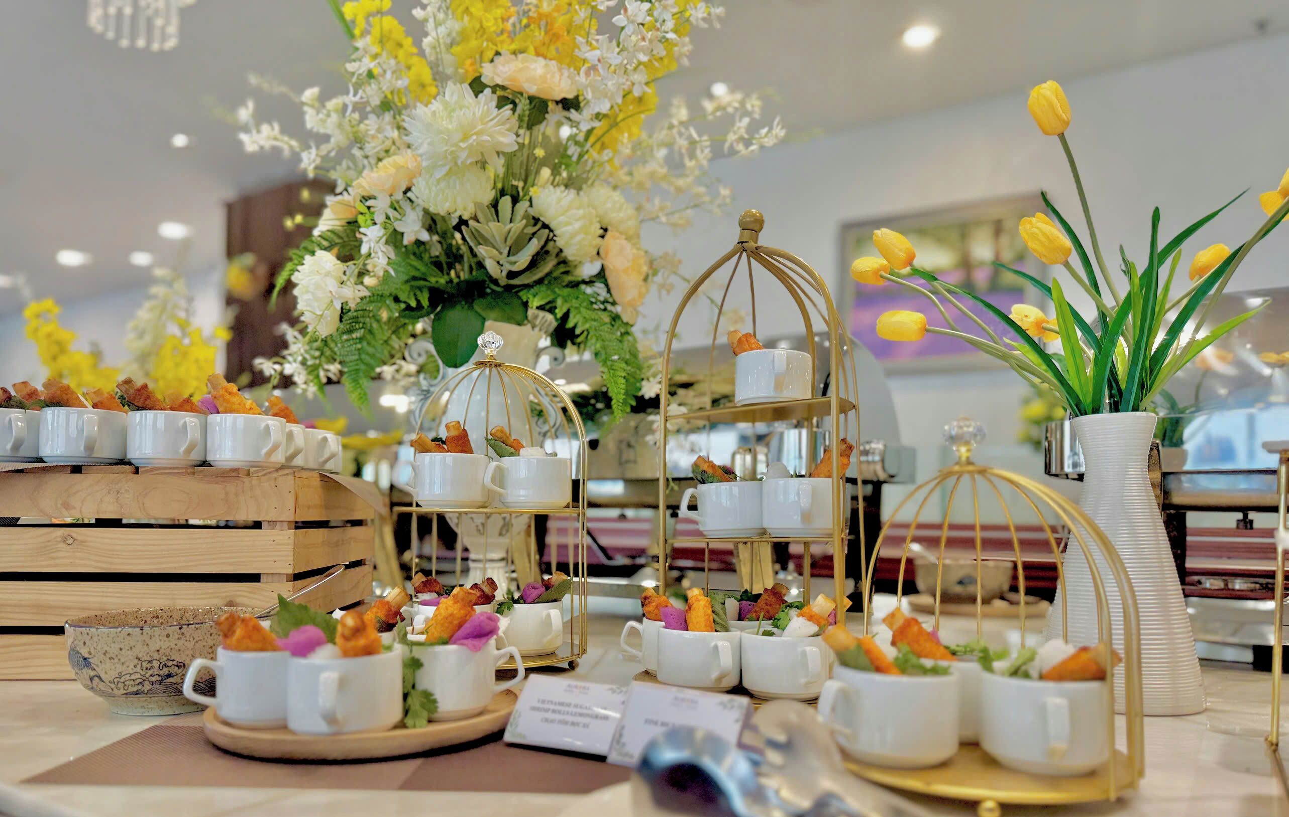 Una mesa dispuesta con tazas y platitos blancos, un centro de mesa floral y una planta en maceta.