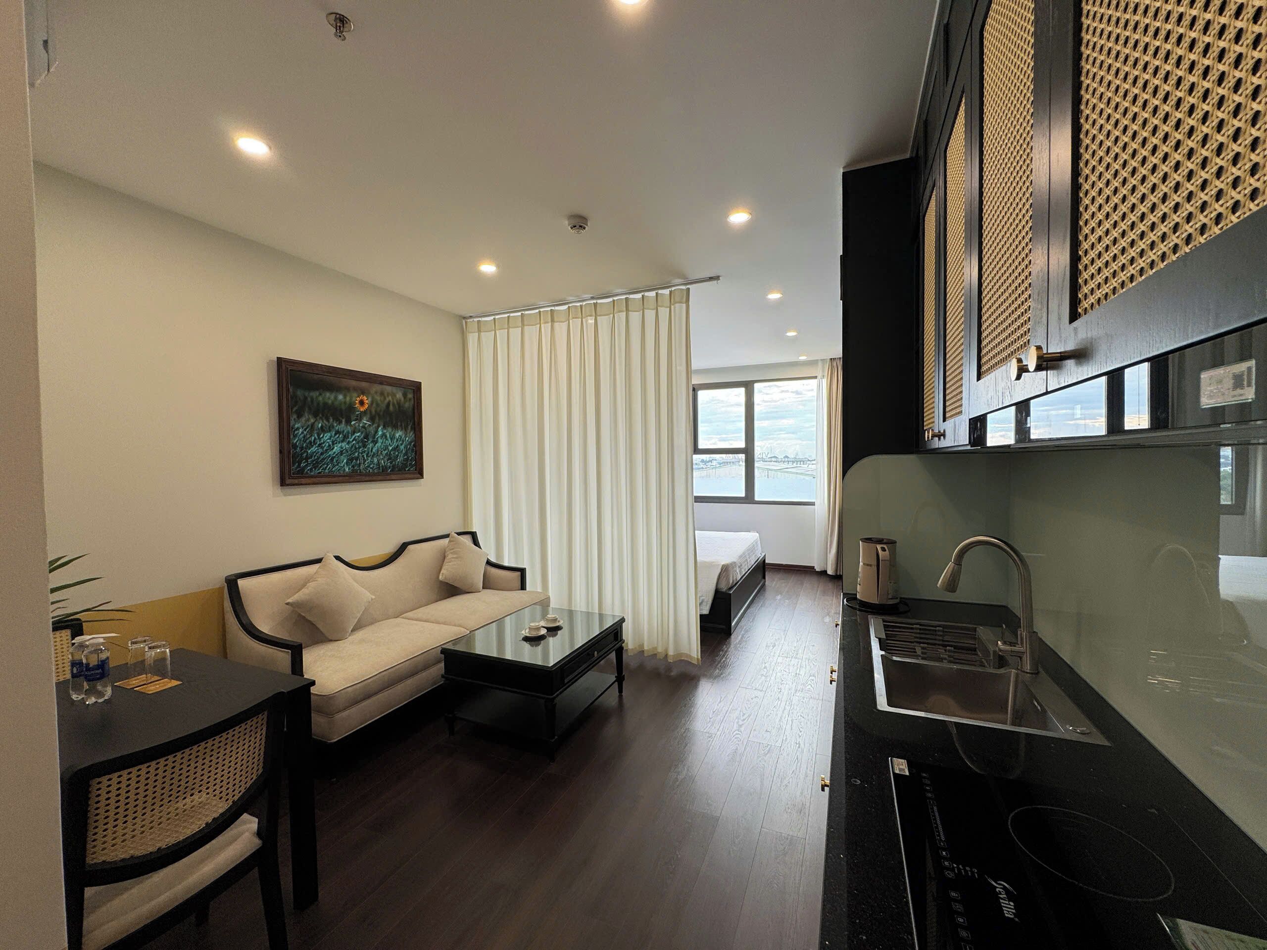 A modern hotel room with a sofa, a small dining table, a kitchen area with a sink and stove, and a large window offering a view of the city.