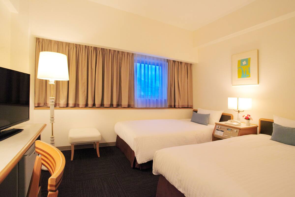 A hotel room with two beds, a desk, a TV, and a window with curtains.