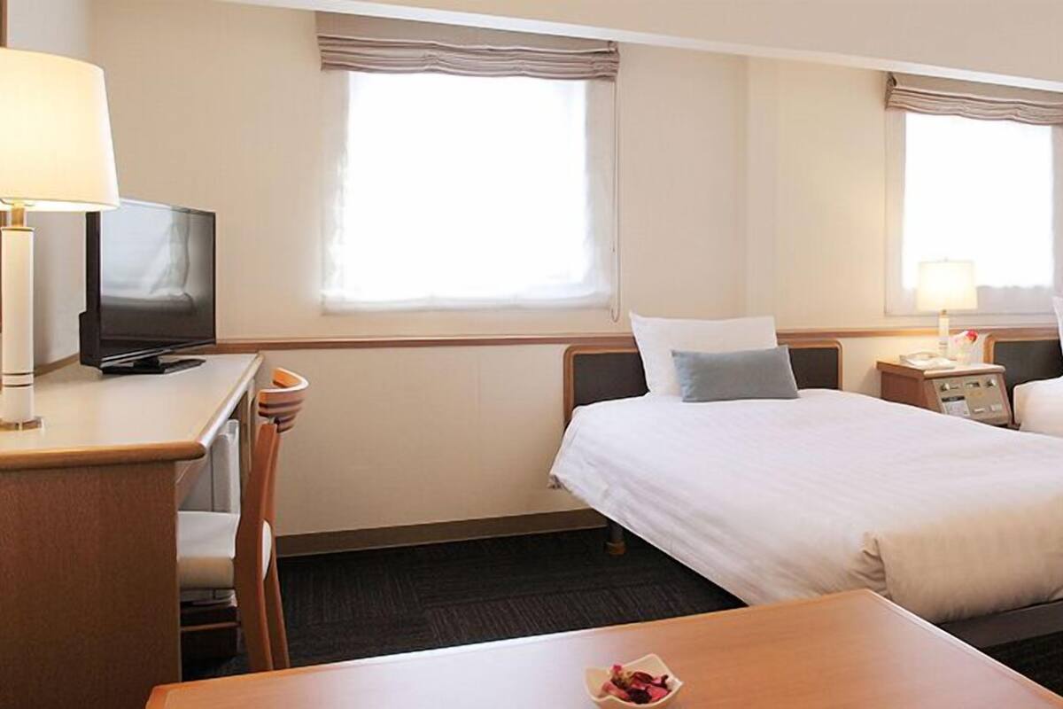 A hotel room with two beds, a desk with a television, a bedside table with a lamp, and a small table with a bowl of fruit.