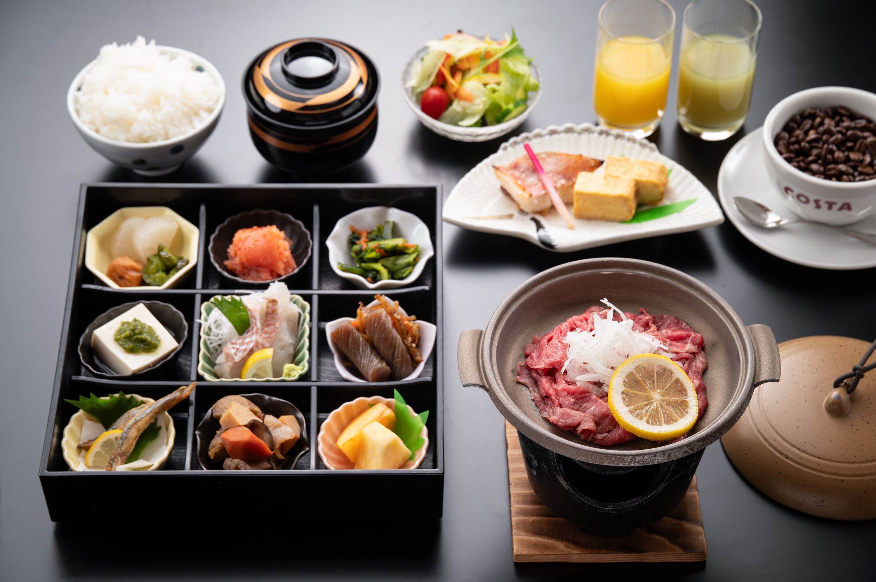 A variety of food items and drinks on a table, including rice, a pot of raw meat, and a bento box with multiple compartments.