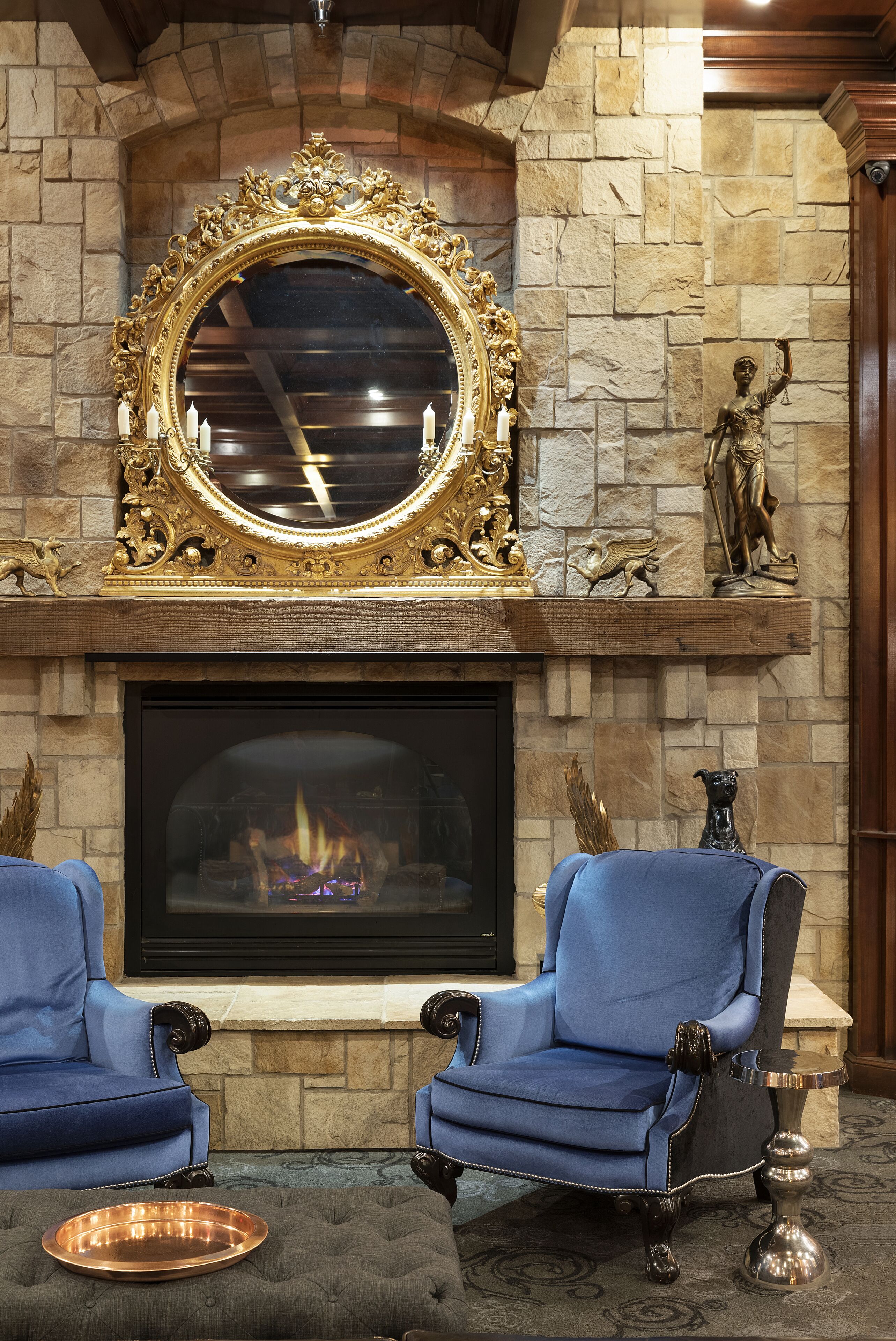 A fireplace with a stone mantle, two blue armchairs, and a decorative mirror above it.