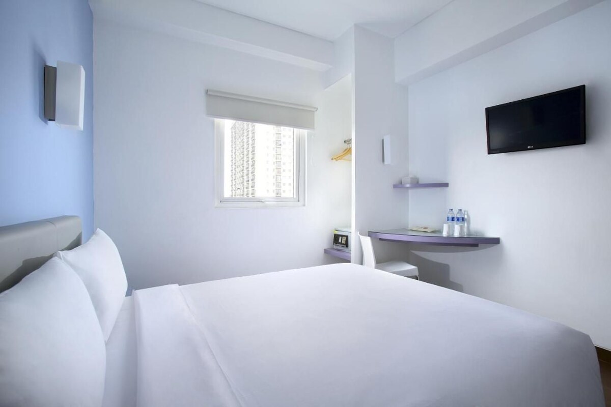 A hotel room with a bed, a desk, a chair, a TV, a window with curtains, and a shelf with water bottles.