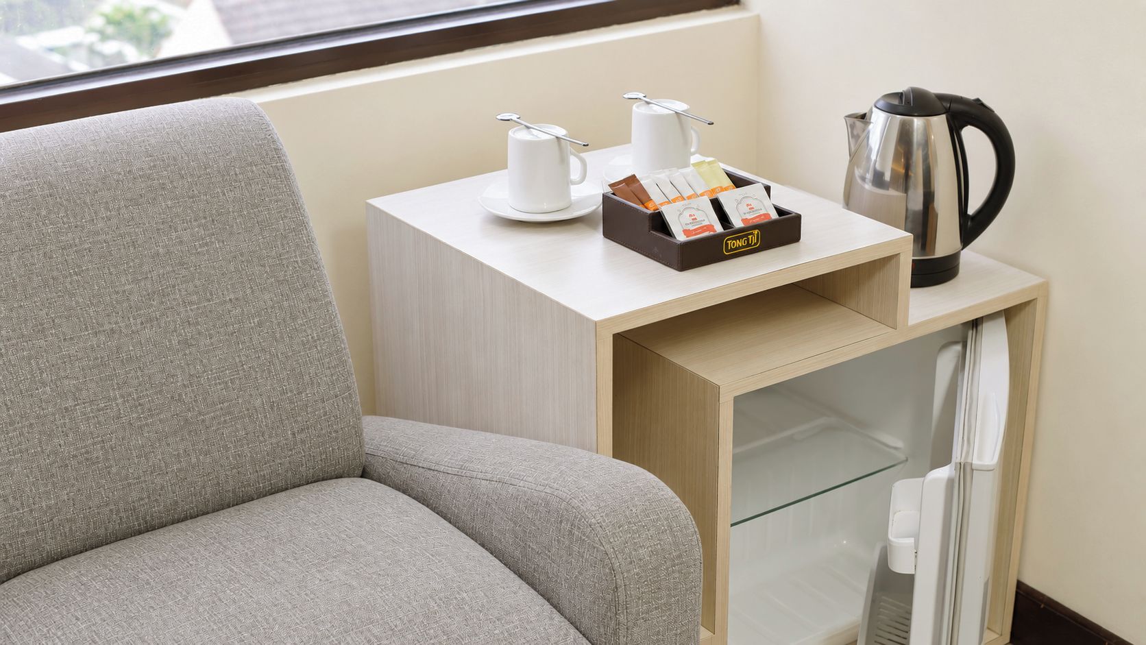 Et hotelværelse med en grå sofa, et lille træ sidebord med kaffemaskine, teposer og elkedel.