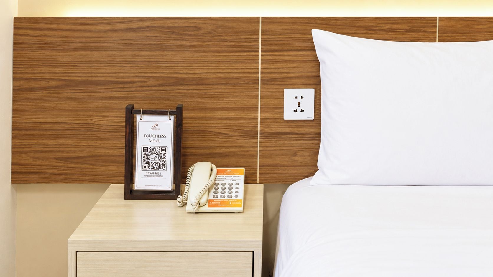A hotel room with a wooden headboard, a bedside table with a phone and a QR code sign, and a white pillow.