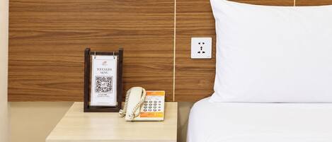 A hotel room with a wooden headboard, a bedside table with a phone and a QR code sign, and a white pillow.