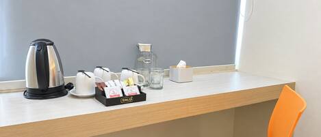 A hotel room with a wooden desk, a kettle, a water pitcher, a tissue box, and a chair.