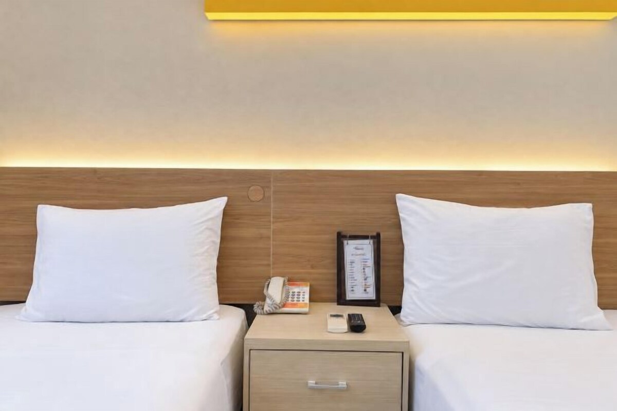Habitación de hotel con dos camas, una mesita de noche con teléfono y un cuadro enmarcado, y una mesita de cabecera con cajón.