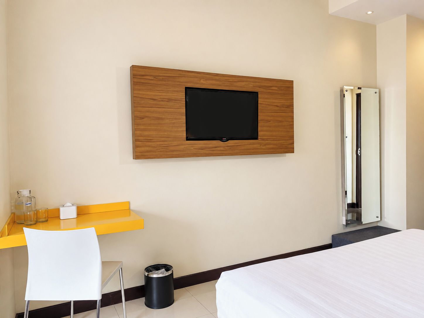 A hotel room with a bed, a desk with a chair, a trash bin, a mirror, and a television mounted on the wall.