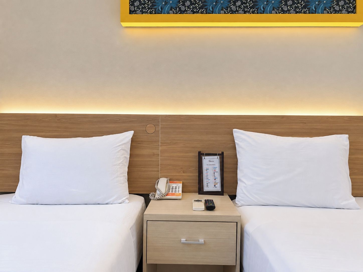 A hotel room with two beds, a bedside table with a phone and remote control, and a wall-mounted TV.