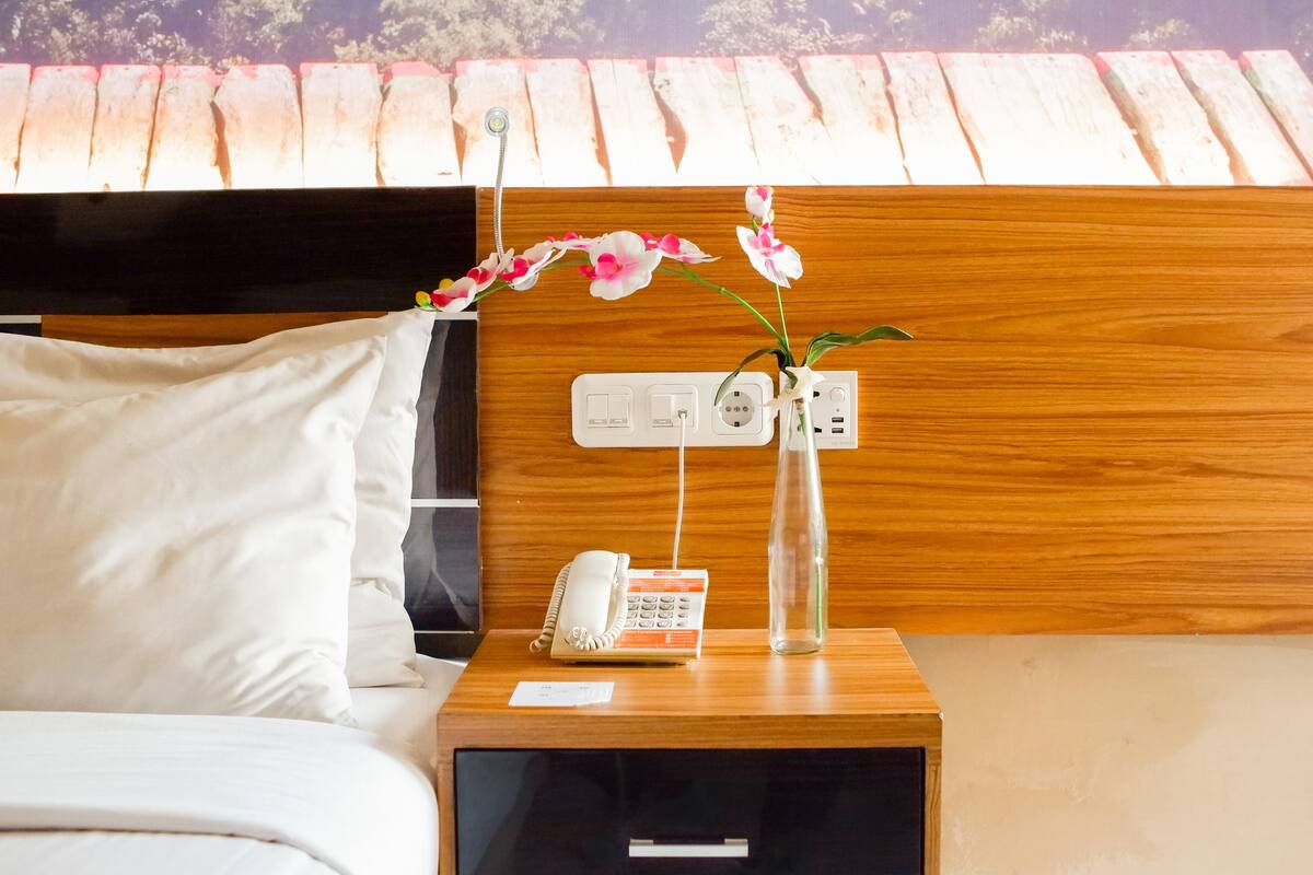 Habitación de hotel con cabecera de madera, mesita de noche con teléfono, un florero con flores y toma de corriente.