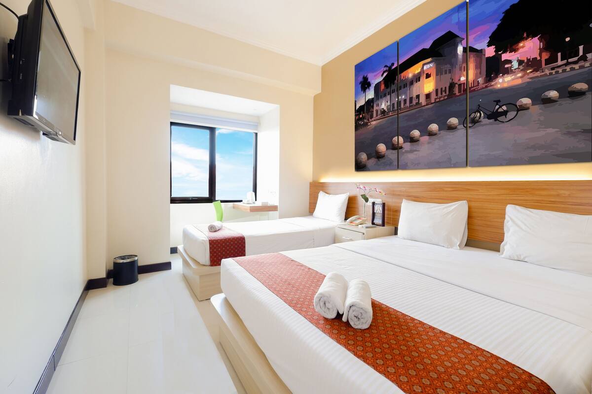 Habitación de hotel con una cama grande, una cama más pequeña, un televisor, una ventana con vista a la calle y un cuadro en la pared.