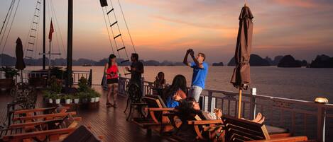 A wooden deck with lounge chairs and tables, overlooking a body of water with distant landforms.