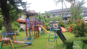 Un parque infantil con diversas estructuras de juego, rodeado de vegetación y edificios al fondo.