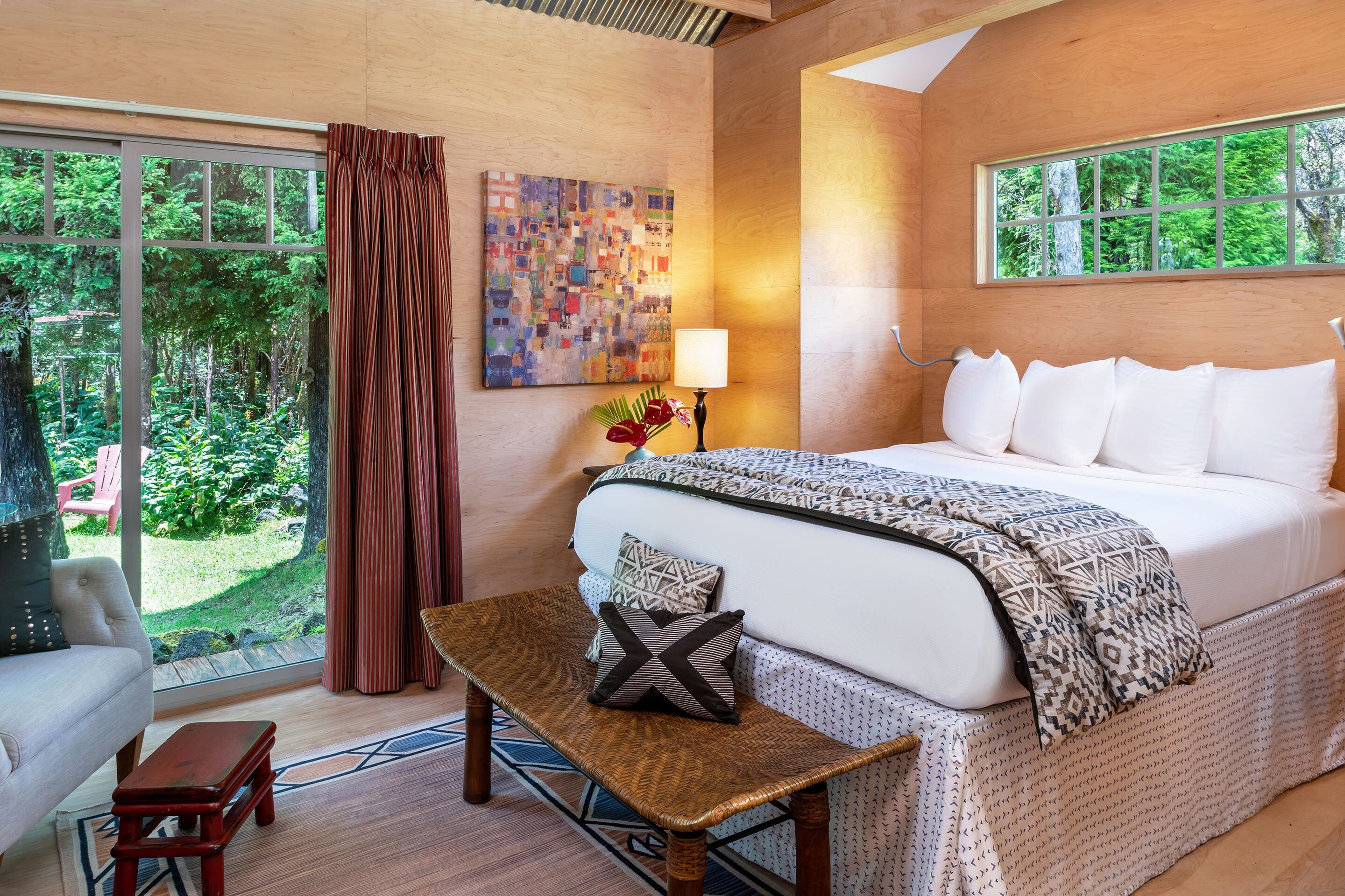 A bedroom with a large bed, a wooden bench, a chair, a window with curtains, and a painting on the wall.