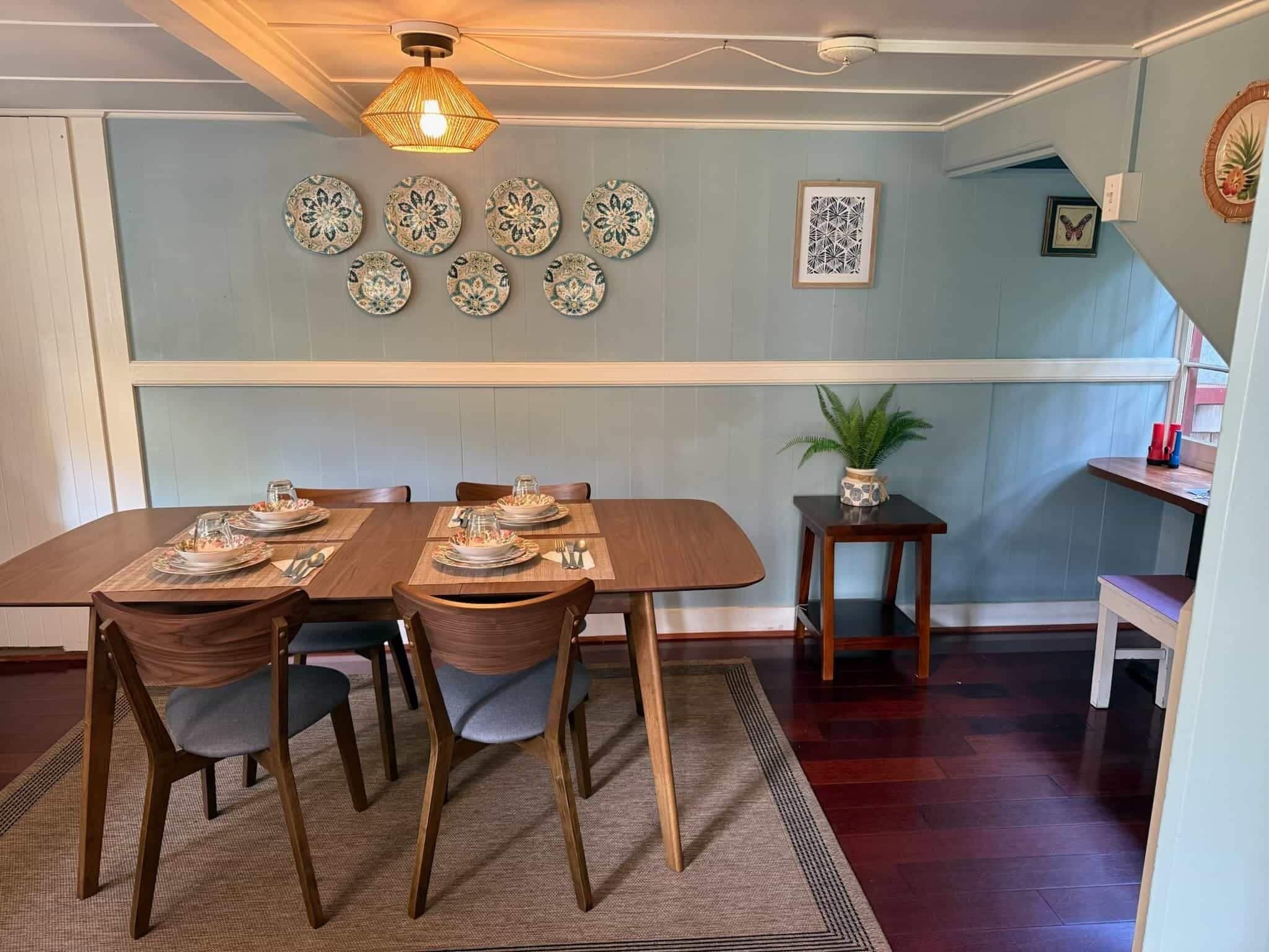 Un comedor con una mesa de madera dispuesta para cuatro personas, rodeada de sillas, una lámpara colgante y platos decorativos en la pared.