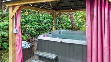 Een hottub onder een houten pergola met roze gordijnen, omgeven door weelderig groen.