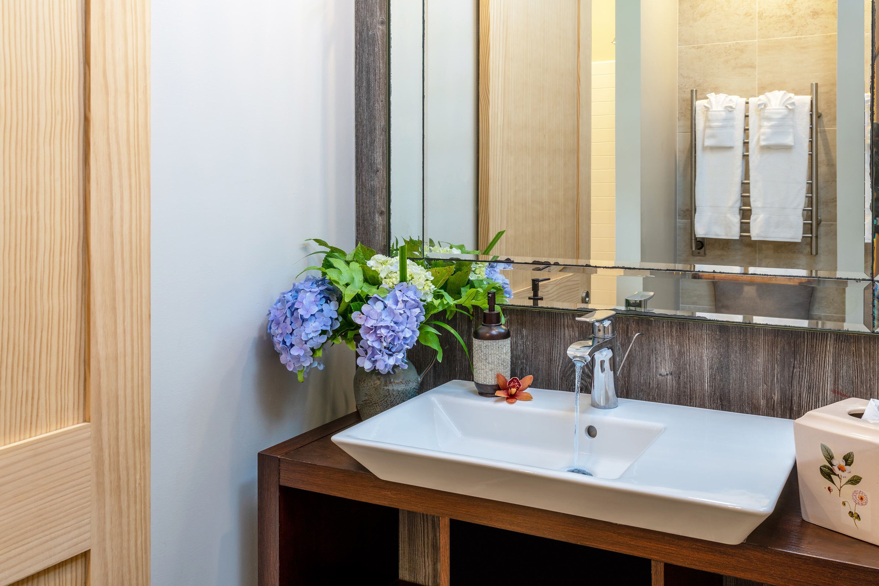 A bathroom with a wooden vanity, a white sink, and a mirror reflecting a towel rack.