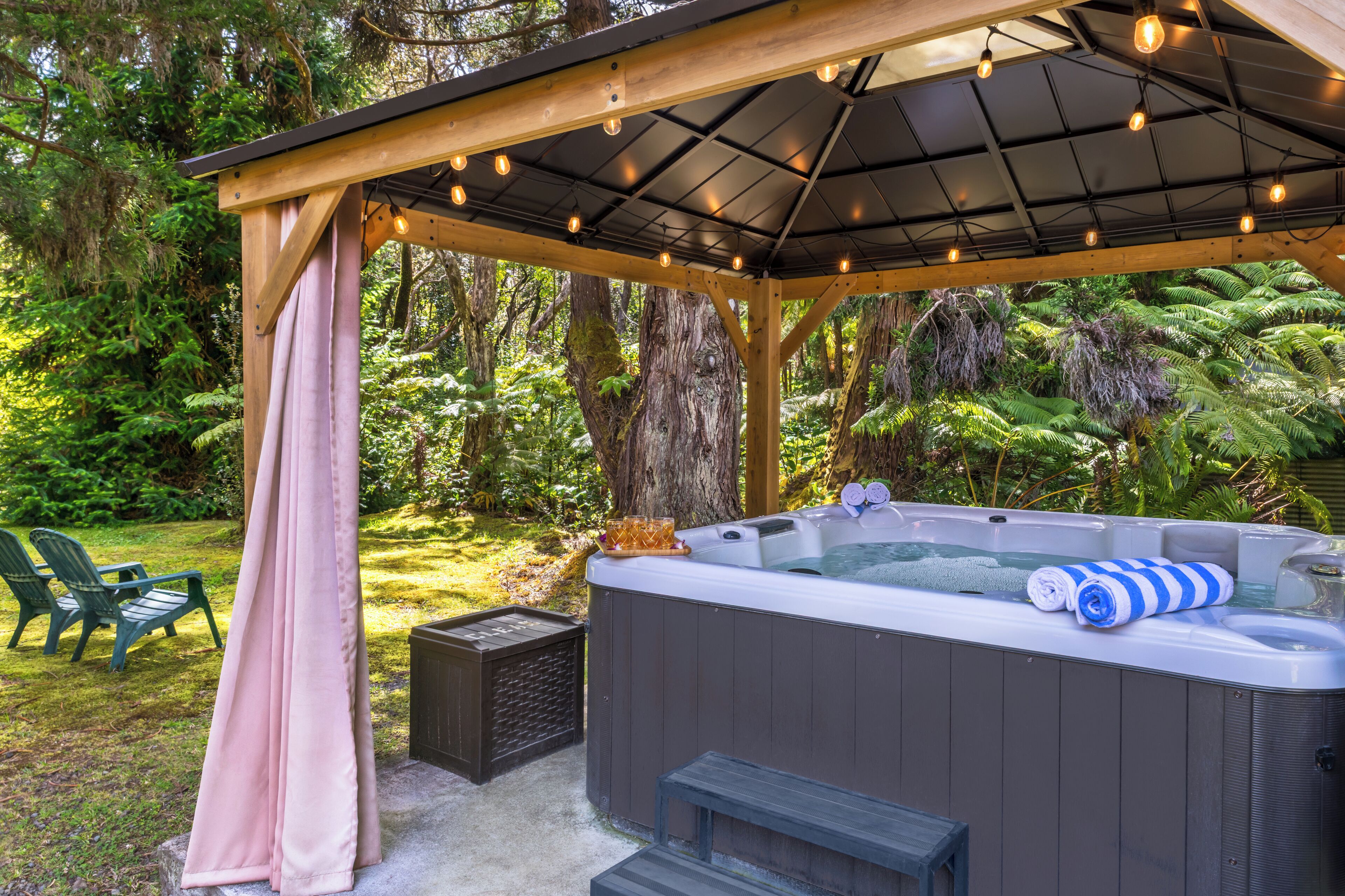 A hot tub under a wooden pergola with string lights, surrounded by lush greenery.