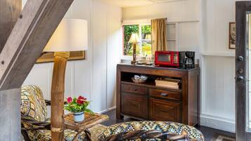 Una habitación con una silla de madera, un otoman con estampado, una mesita con un jarrón de flores, un mueble de madera con microondas y una puerta con panel de vidrio.