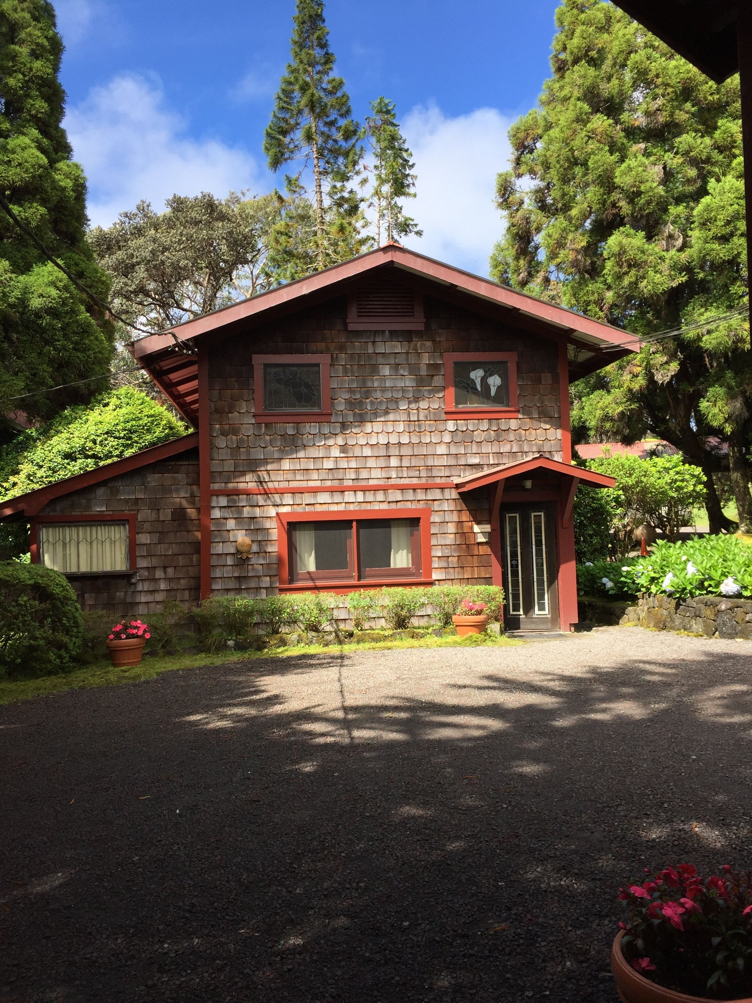A two-story wooden house with a red roof, surrounded by lush greenery and trees.