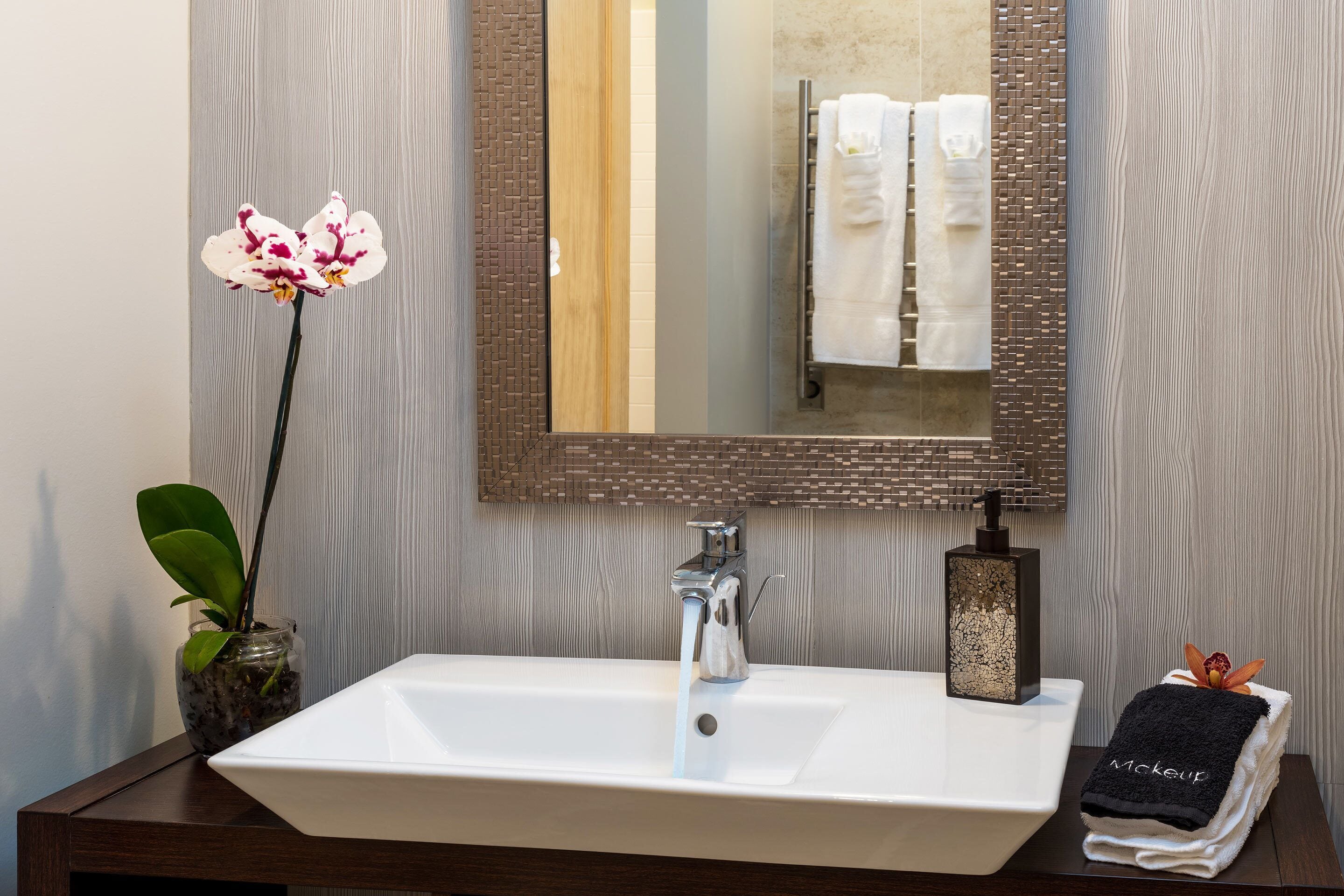 Un baño con lavamanos blanco, espejo, orquídea en maceta y toallero con toallas.