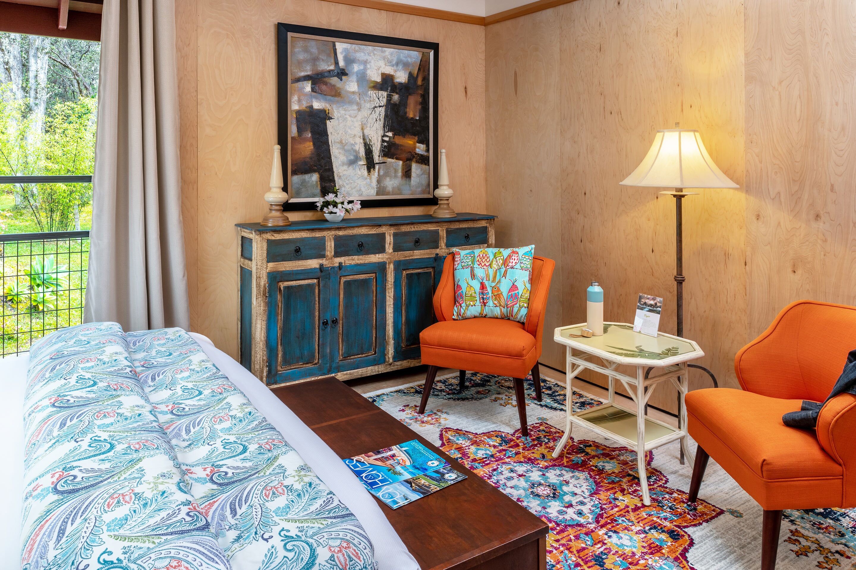 Habitación de hotel con una cama, dos sillones naranjas, una mesita, una lámpara, un armario y un cuadro en la pared.