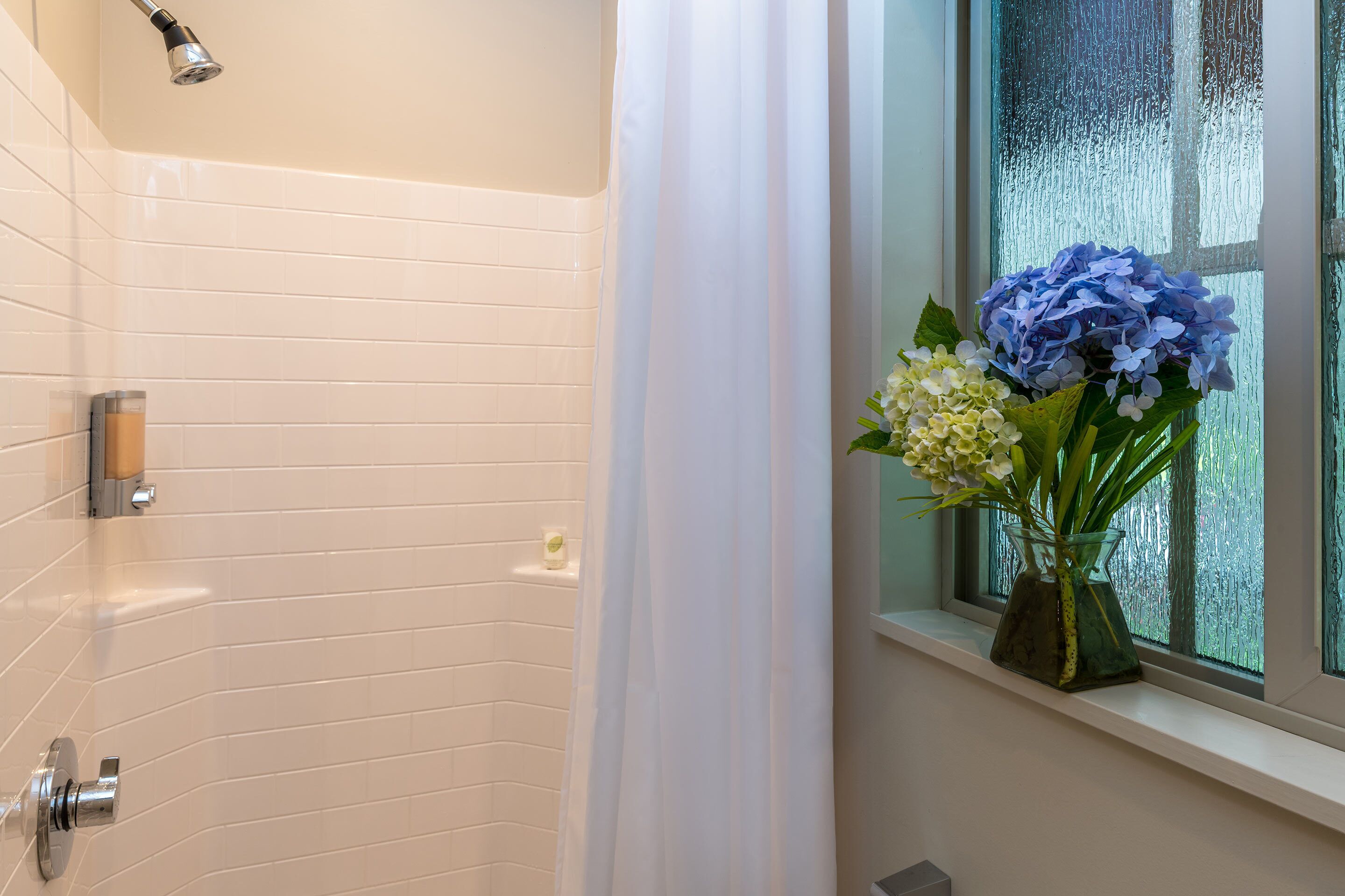 A bathroom with white tiled walls, a shower, a window with a view, and a vase with flowers on the windowsill.
