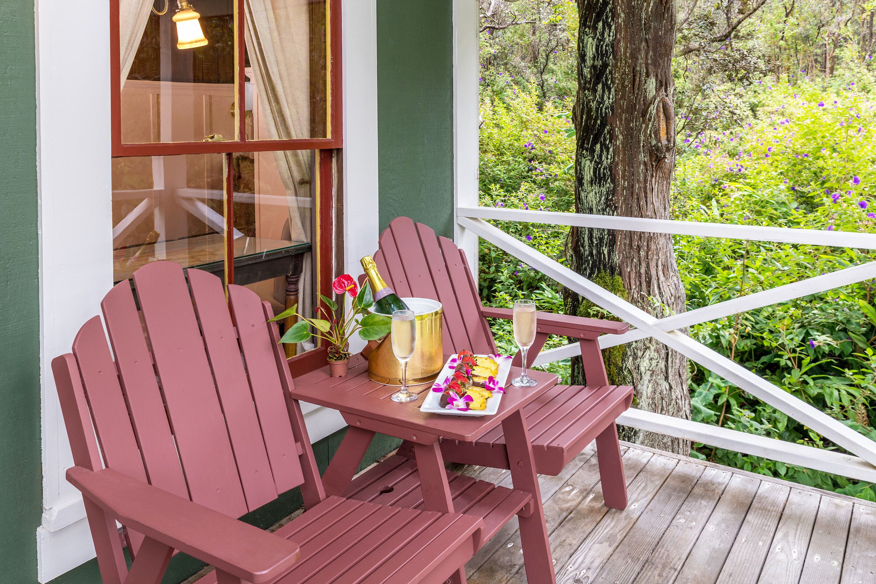 A wooden deck with two Adirondack chairs, a table with a bottle of champagne, and a tray with flowers.