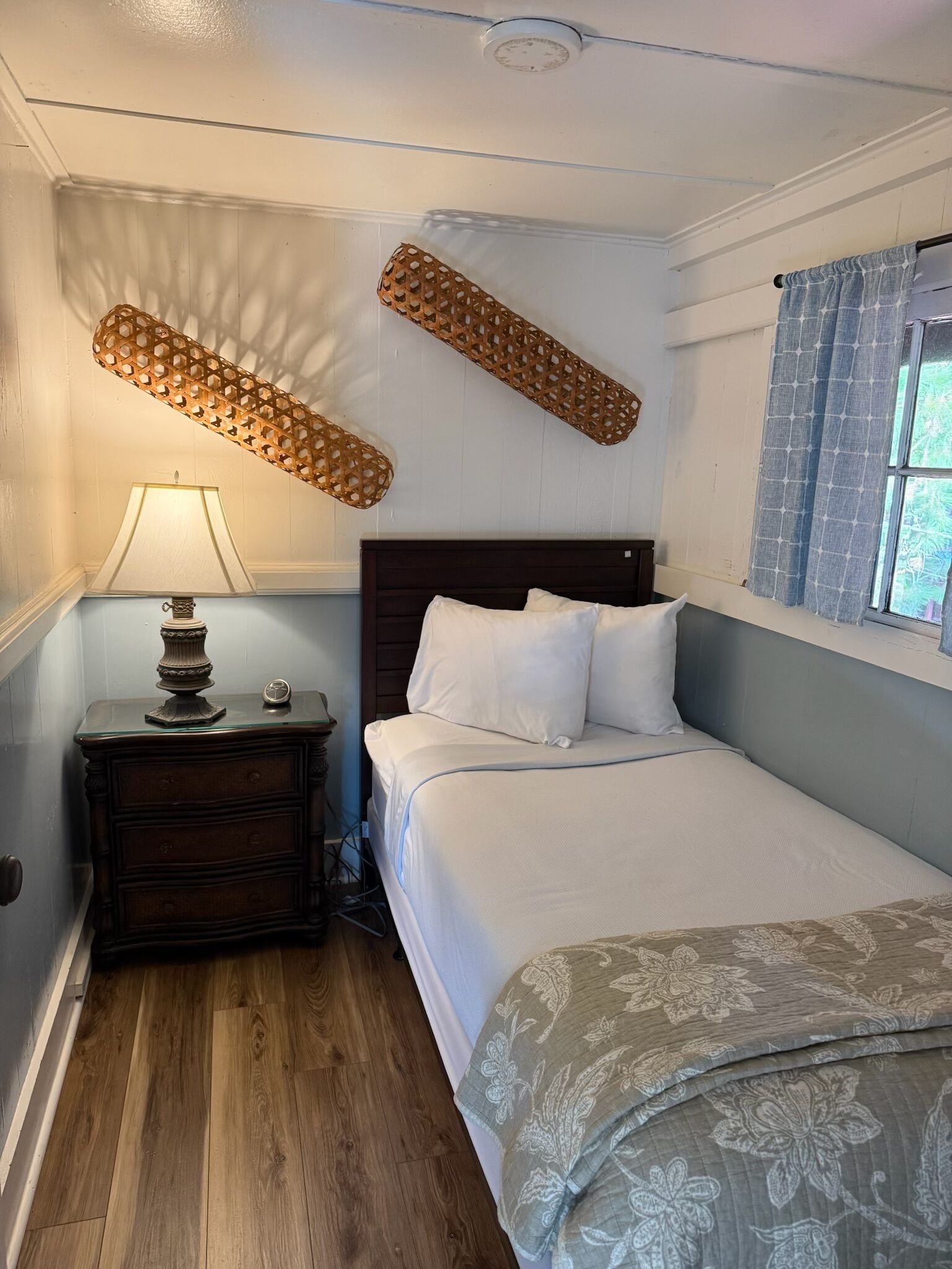 Une chambre à coucher avec une table de chevet en bois, une lampe, un lit avec une literie blanche et une fenêtre avec des rideaux.