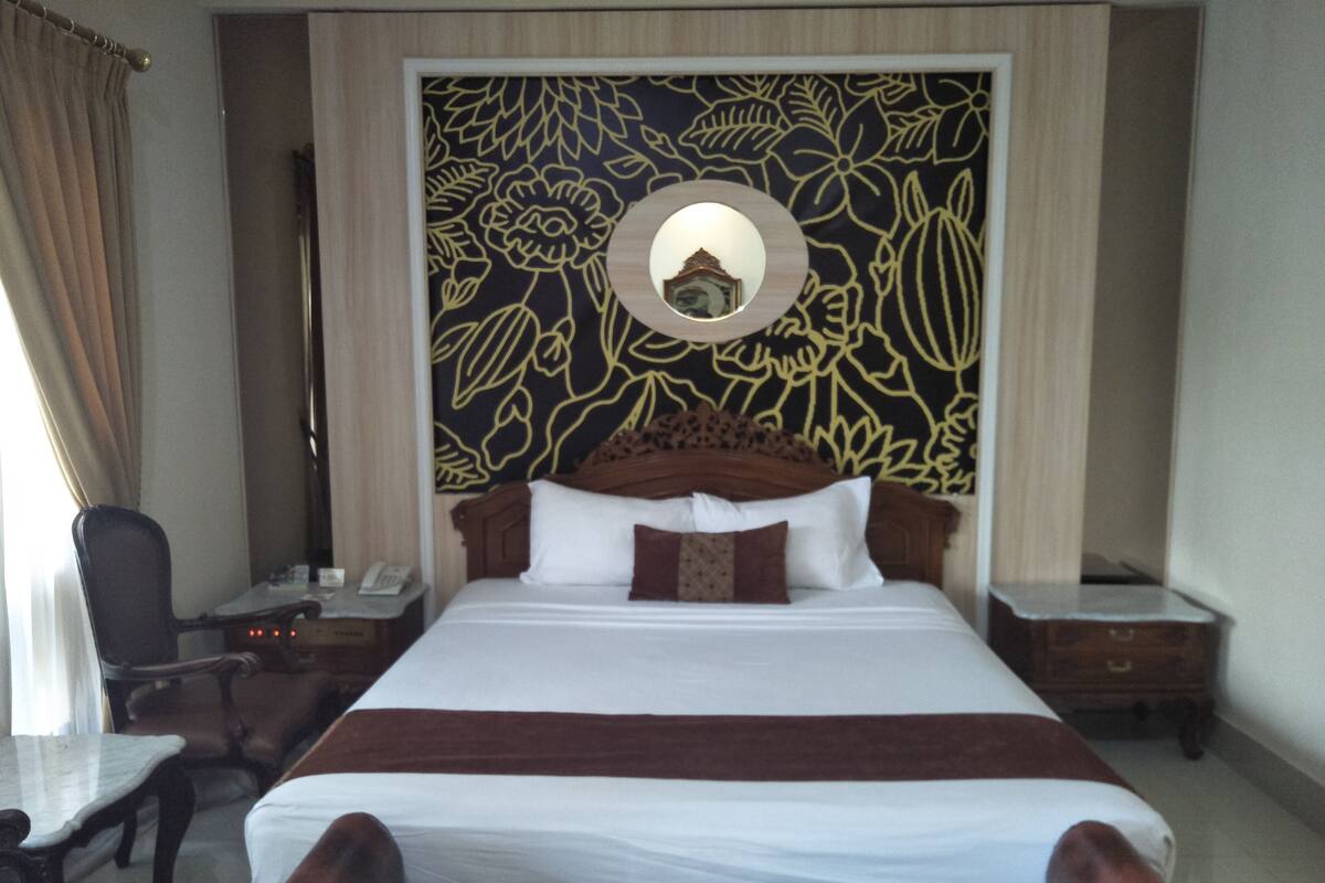 A hotel room with a large bed, a wooden headboard, a nightstand with a lamp, a desk with a chair, and a wall with a floral pattern.