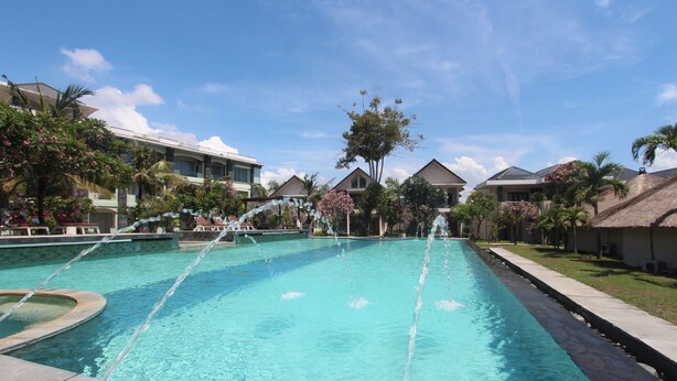 A swimming pool with clear blue water, surrounded by palm trees and a building with a thatched roof. - Alit Beach Resort and Villas (Denpasar)